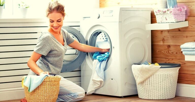 a woman kneeling while loading freshly washed clothes, including a light blue tie-dye shirt, into a dryer for proper tie-dye shirt care