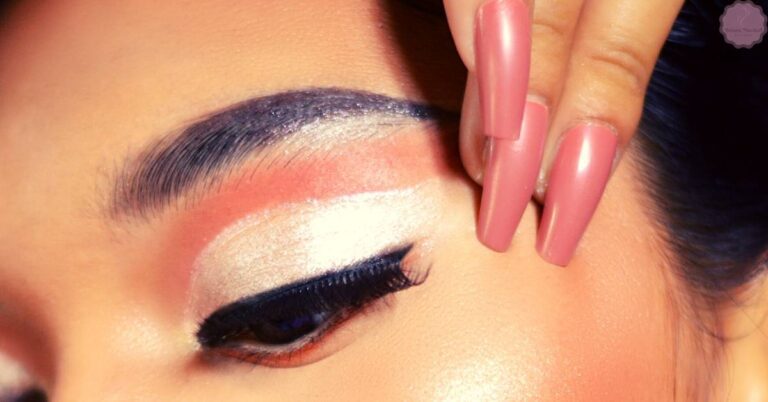 a close-up of a person’s eye with dramatic cool-toned eyeliner and shimmery eyeshadow application, accented by long lashes and a hand holding a pink nail polish brush.