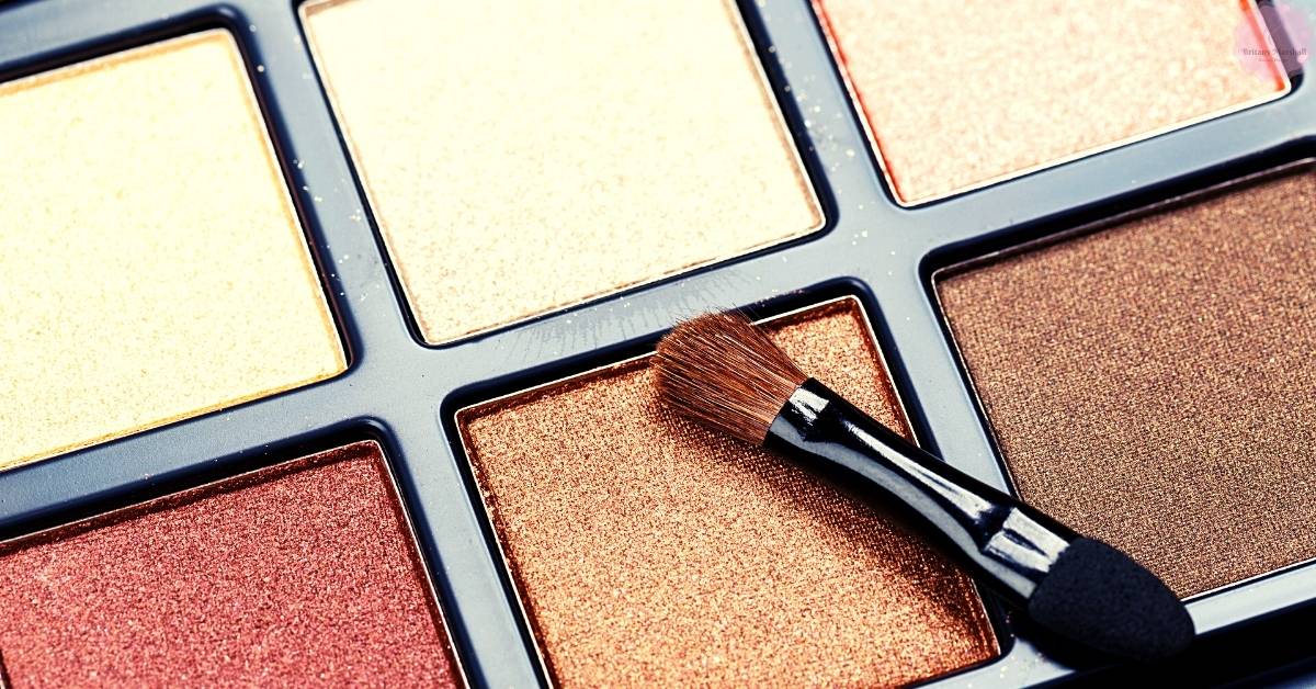 a close-up of an open eyeshadow palette with a single shimmery gold shade being applied using a fluffy brush