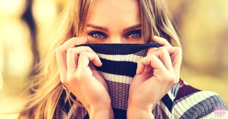 a person covering their face with a striped scarf, revealing striking blue eyes and natural makeup highlighting their expressive gaze.