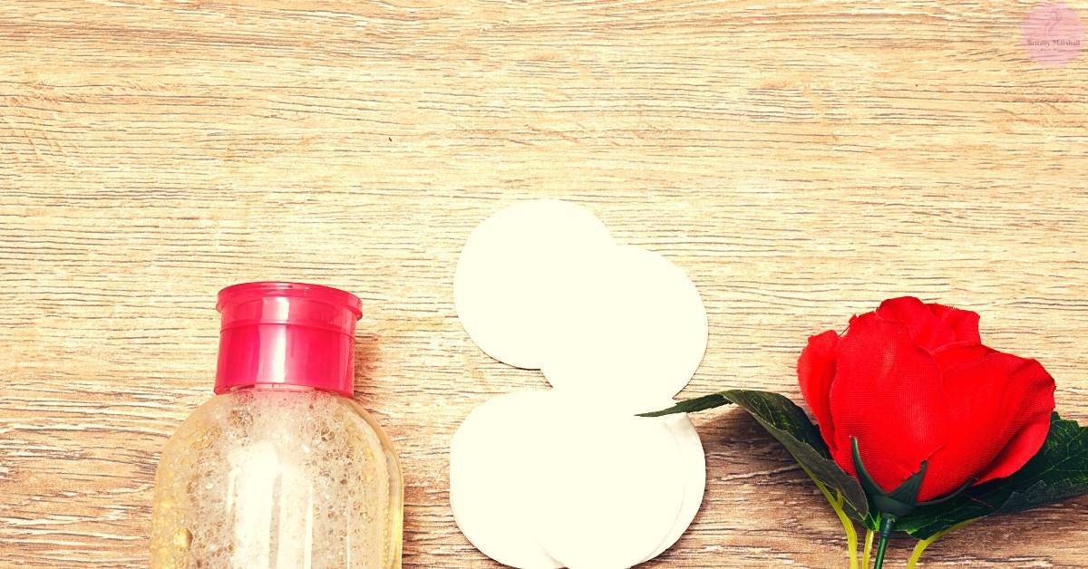 a red bottle of oil-based makeup remover beside a white cotton pad and a red rose on a textured wooden surface