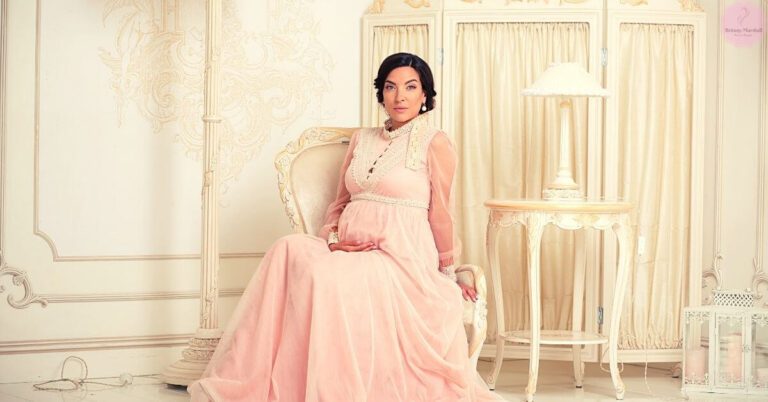 a woman seated elegantly in a light pink, long-sleeve maternity gown with lace detailing, posing in a luxurious room.