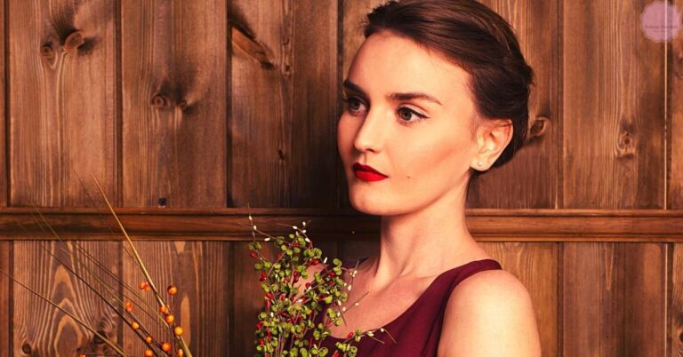 a woman in an elegant burgundy dress holding greenery, with sophisticated makeup featuring bold red lips and a natural look.