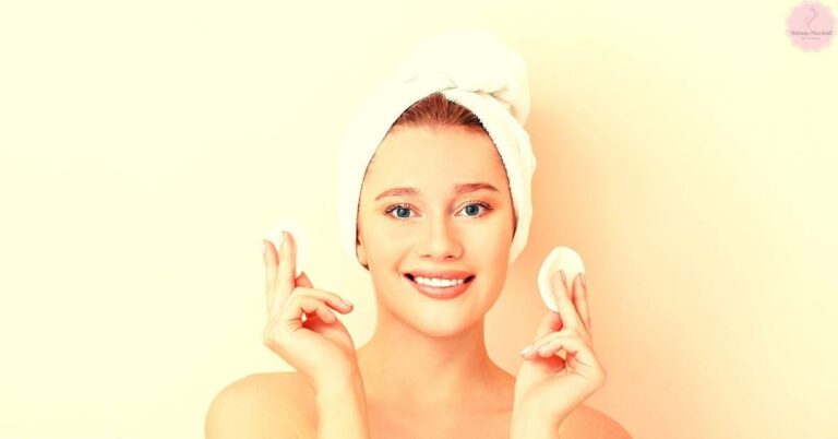 a smiling person with warm peach undertone skin wearing a towel turban, holding a makeup sponge and gesturing peacefully.
