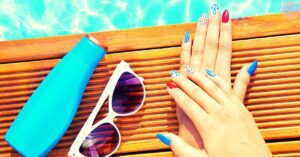a hand displaying brightly colored nail polish with a peach and red shade, white and blue accents, next to a blue nail dryout topcoat and white sunglasses on a wooden bench