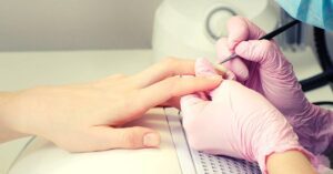 a person receiving a gel nail polish removal using an acetone-soaked pad while wearing protective gloves.