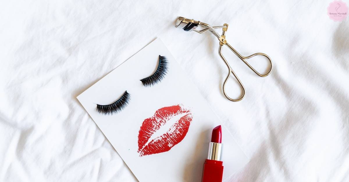 a set of false eyelashes, a gold-tipped eyelash curler, and a red lipstick with a printed lip stamp on a white background