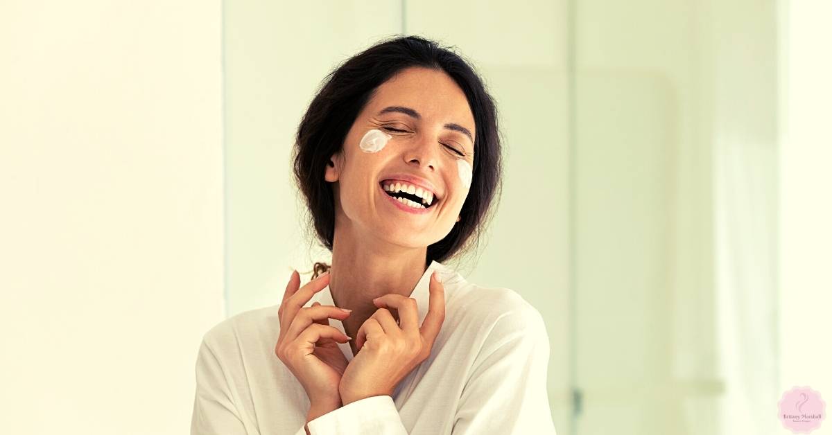 a woman smiling with eyes closed, applying a light facial moisturizer or skincare product near her eyes