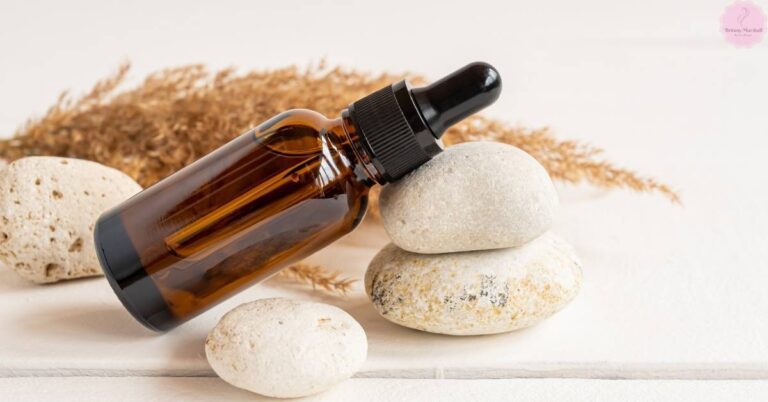 a brown glass dropper bottle with black cap placed beside smooth pumice stones and natural wheat stalks on a light surface