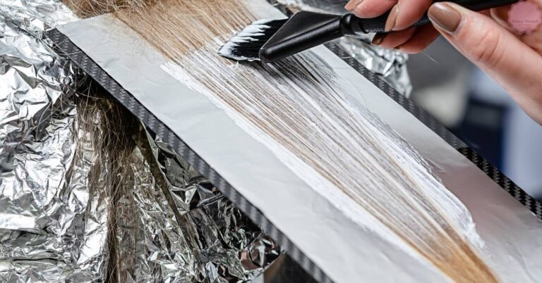 a close-up of hands applying aluminum foil-wrapped hair bleach strips to damp hair for a lightening treatment