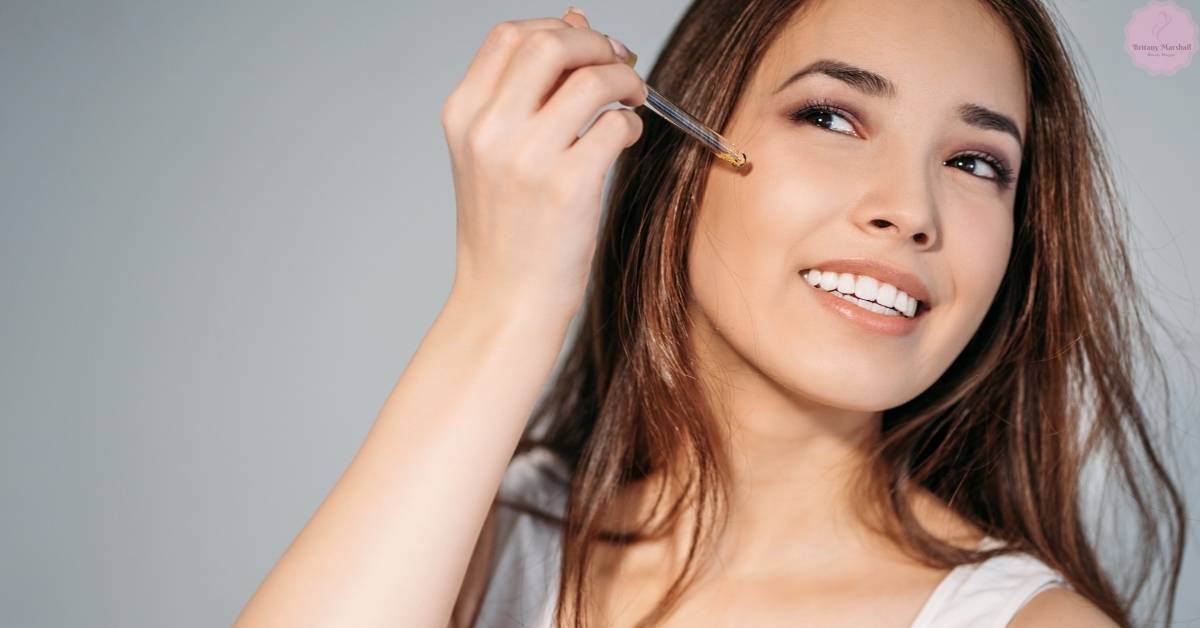 a woman smiling while applying a skincare serum near her eye area with a precision dropper.