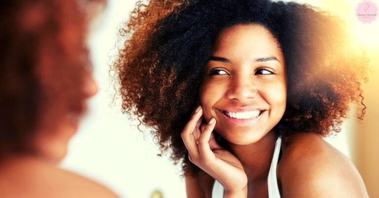a smiling woman with voluminous curly hair touching her face, radiating joy in natural sunlight