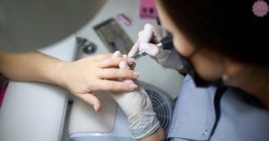 a person receiving nail maintenance with a technician applying acetone to gel nails using a cuticle tool