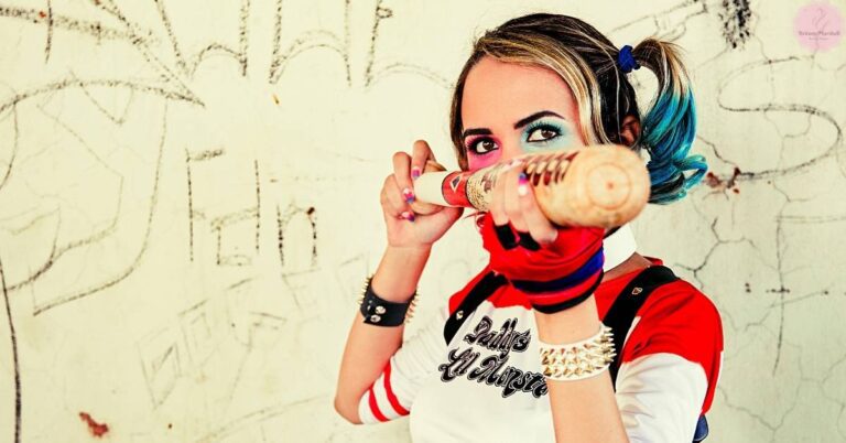 a person demonstrating a bold Harley Quinn-inspired makeup look with dramatic red and black face paint, blue hair, and a matching costume.