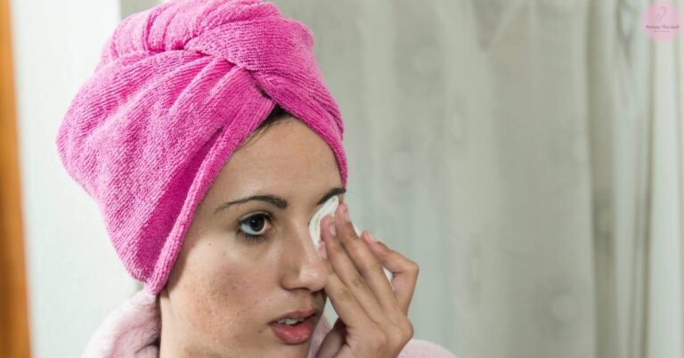 a person wearing a pink towel turban gently cleansing their eye area with a cotton pad for delicate eye skin care