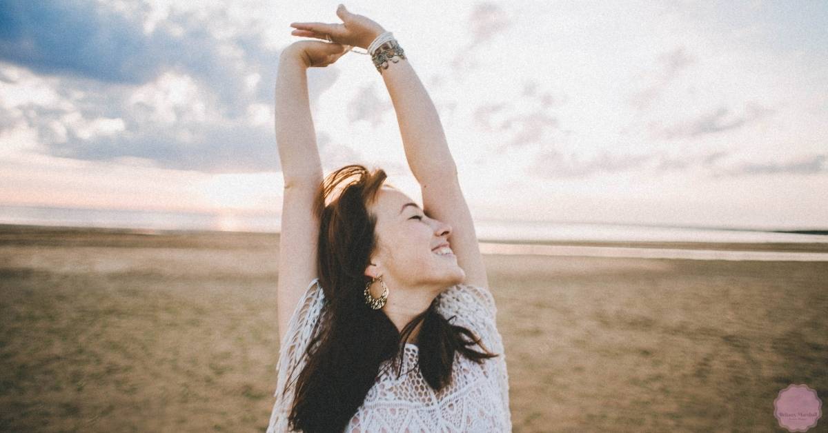 a person stretching arms upward outdoors, smiling with natural light, embodying confidence and self-care.