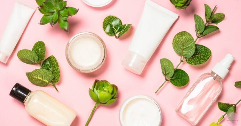 a collection of natural skincare products and green botanicals on a pink background, including lotions, serums, and leaves