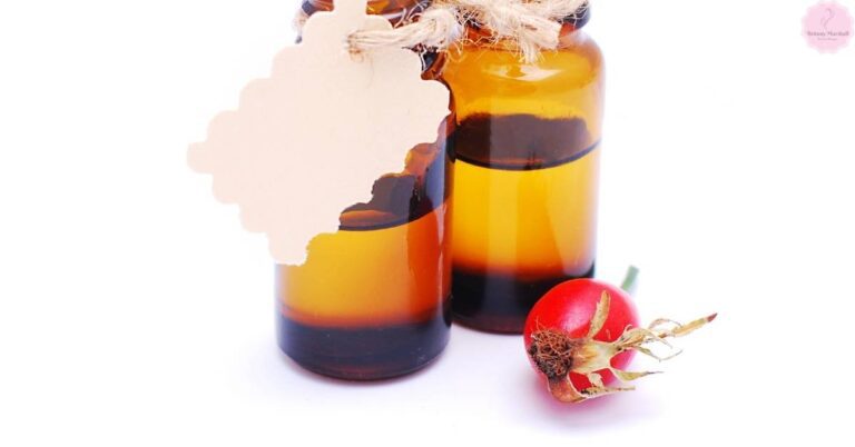 Two amber glass jars with dark liquid rosehip oil, tied with twine, beside a dried rosehip fruit on a plain background.