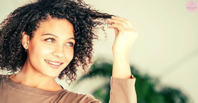 a smiling woman with curly hair lifting a strand to show sun-damaged or lightened tips, highlighting potential hair texture changes.