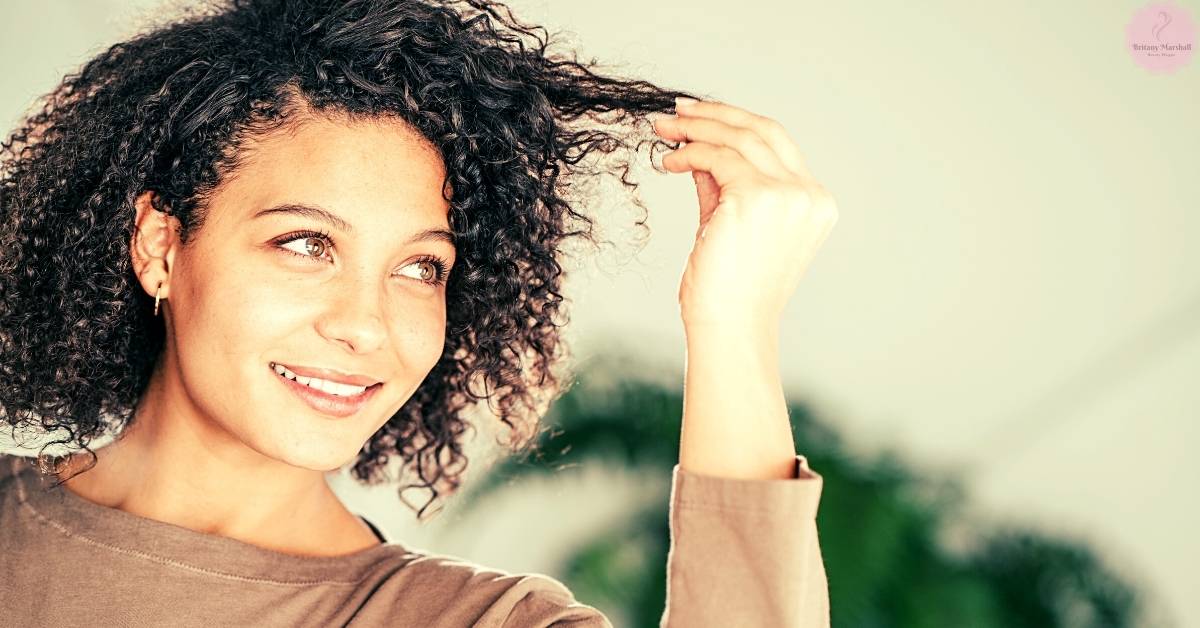a smiling woman with curly hair lifting a strand to show sun-damaged or lightened tips, highlighting potential hair texture changes.