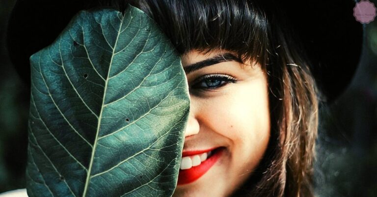 a smiling person partially hiding behind a large green leaf, showcasing vibrant red lipstick.
