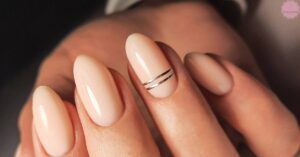a hand showcasing short almond-shaped nails with a sleek beige polish and subtle metallic stripe detail on one nail