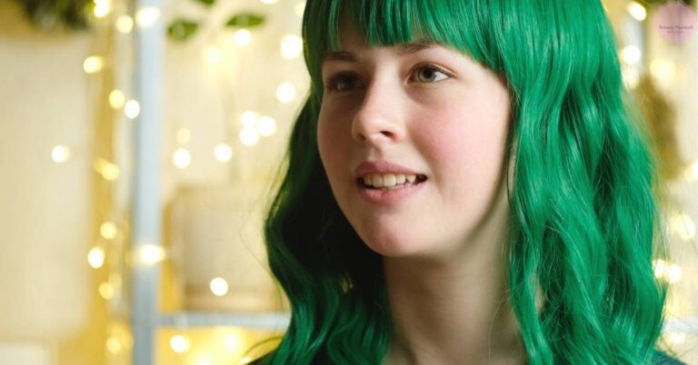 a person with vibrant green hair and bangs smiling softly in a warm, bokeh-lit indoor setting