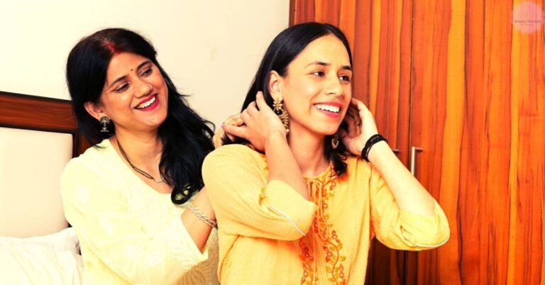 two smiling women adjusting each other’s earrings in traditional attire, indoors.