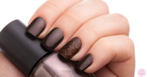 a close-up of a hand holding a dark brown nail polish bottle with four dark brown acrylic nails and one nail featuring brown glitter nail art.