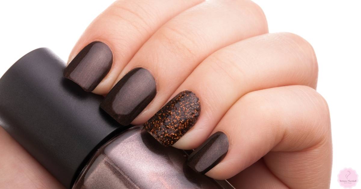a close-up of a hand holding a dark brown nail polish bottle with four dark brown acrylic nails and one nail featuring brown glitter nail art.