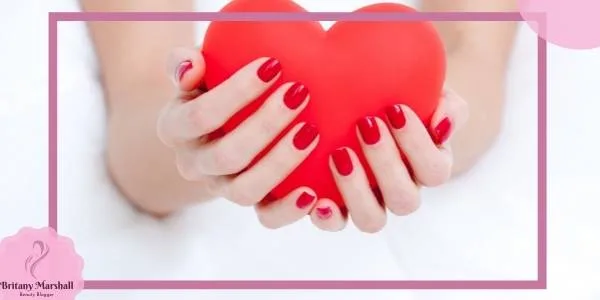 a pair of hands gently holding a red heart-shaped object with neatly painted light pink and mauve nail polish