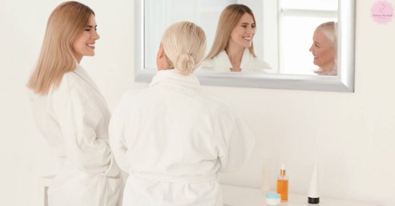 a woman smiling while observing another person’s reflection in a mirror during a skincare routine with a discoloration correcting serum on a vanity.