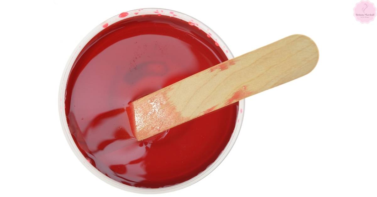 a clear bowl containing red manzilian wax with a wooden stick for application