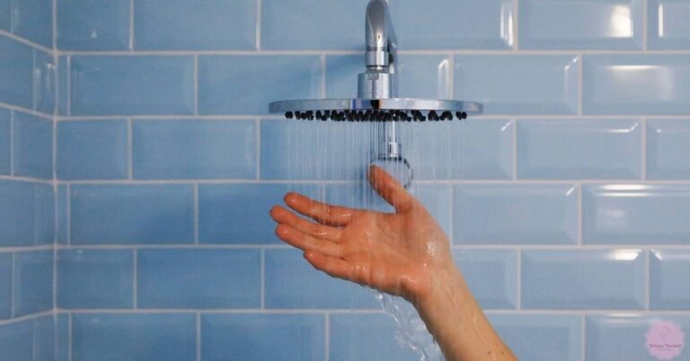 a hand testing warm shower water from a rainfall showerhead against light blue tiled wall for skin preparation