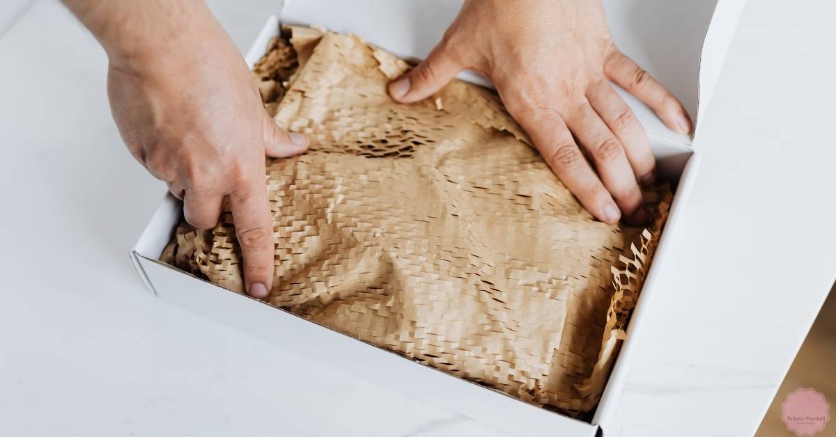 a pair of hands carefully placing a shredded paper filler into a white press-on nails display box