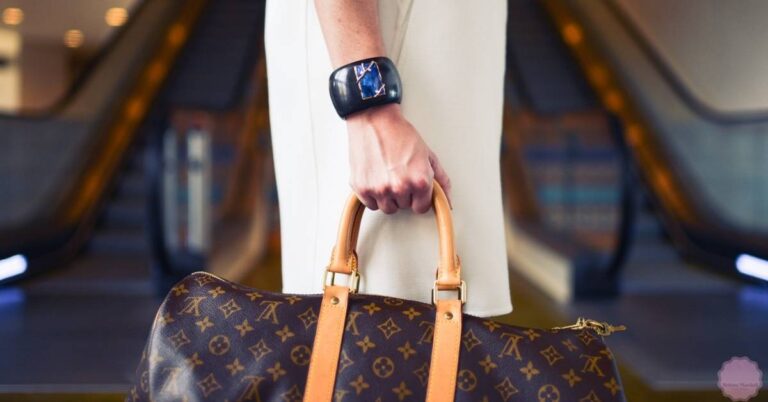 a person holding a Louis Vuitton monogram handbag with tan leather handles and straps, wearing a distinctive dark wristwatch.