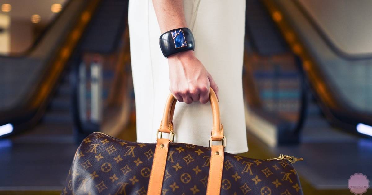 a person holding a Louis Vuitton monogram handbag with tan leather handles and straps, wearing a distinctive dark wristwatch.