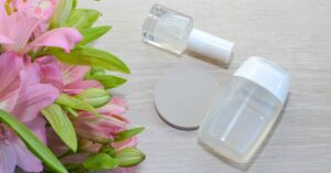 two clear bottles of acetone-based nail polish remover with applicator brushes beside pink lilies on a light wooden surface