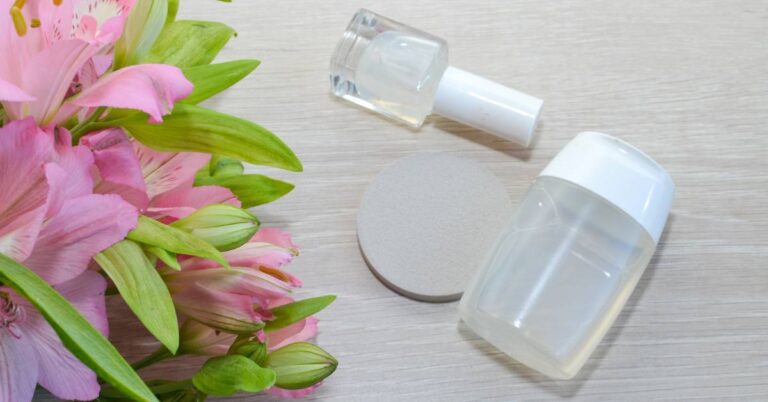 two clear bottles of acetone-based nail polish remover with applicator brushes beside pink lilies on a light wooden surface