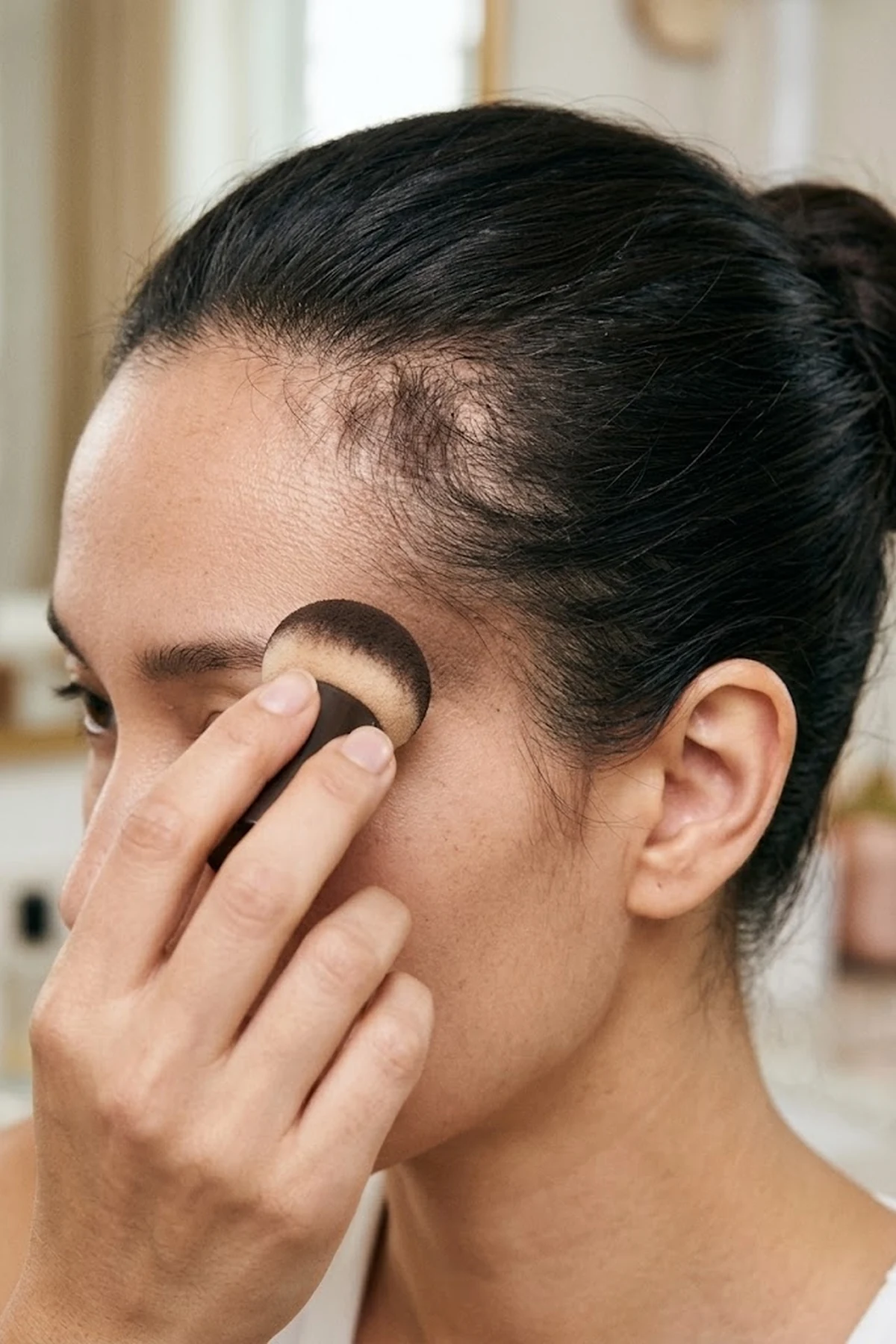 Close-up beauty photo of a mushroom-tip cushion sponge stamping espresso brown hair makeup onto sparse temple corners on pulled-back hair (AI Generated Image)