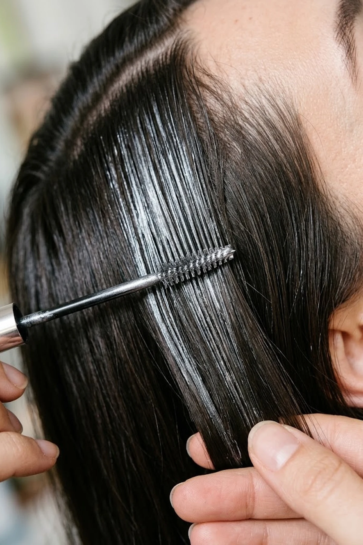 Editorial macro photo of a metallic silver hair mascara spoolie wand brushing chrome grey streaks onto sleek dark espresso hair (AI Generated Image)